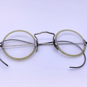 May include: Antique spectacles featuring round, clear lenses encased in pale yellow, translucent circular rims. The frame is constructed from thin, dark metallic wire, including a high-arched bridge and curved temple arms designed to hook behind the ears.