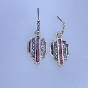 May include: A pair of silver-toned Art Deco style drop earrings. Each earring features a rectangular, red-colored center stone, framed by small, dark gray stones. The earrings have a post and dangle design.