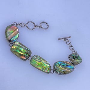 May include: A silver-toned link bracelet featuring five polished abalone shell segments. The iridescent shell pieces display vibrant green, blue, and gold hues. The bracelet is secured with a silver-toned toggle clasp and includes a short chain extension.