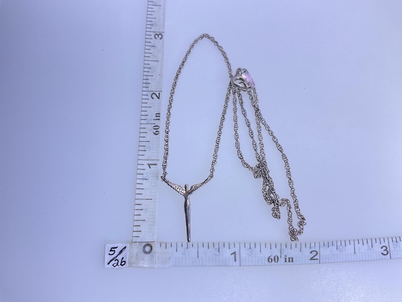 K&ouml;nnte beinhalten: Silberfarbene Halskette mit einem Anh&auml;nger, der eine Figur mit ausgestreckten Armen darstellt. Die Kette ist filigran. Die Halskette ist etwa 5 cm lang, wie auf dem Lineal im Bild angegeben.