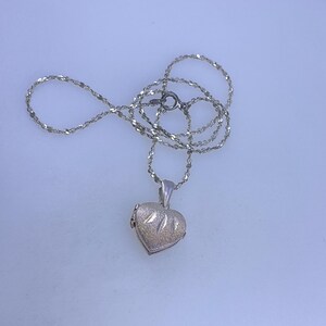 May include: A silver necklace with a heart-shaped pendant. The pendant has a textured surface and a small loop for the chain. The chain is delicate and has a twisted design. The necklace is displayed on a white surface.
