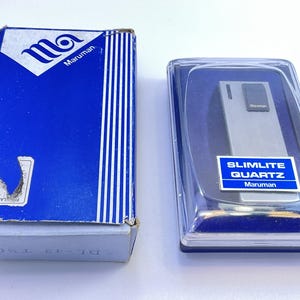 May include: A vintage Maruman Slimlite Quartz lighter in its original packaging. The lighter is silver-toned and rectangular, displayed in a clear plastic case. The blue box features the Maruman logo and text. The lighter's case has a blue and white label.
