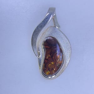 May include: Silver pendant with a teardrop shape. The pendant features a polished, amber-colored stone with gold flecks, set in a textured silver frame. The bail is a simple, curved design.