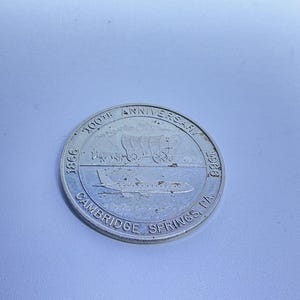 May include: A silver coin commemorating the 100th Anniversary of Cambridge Springs, PA, with the years 1866 and 1966. The coin features a covered wagon and an airplane.