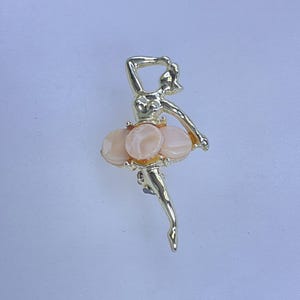May include: A gold-toned ballerina brooch. The dancer is posed mid-leap, with one arm raised. The tutu is composed of three oval, light pink stones. A classic jewelry piece.