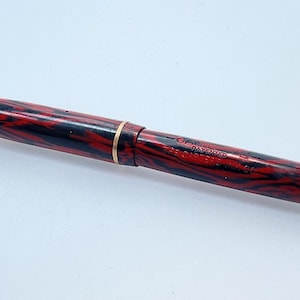 May include: A red and black marbled fountain pen with a gold band and a clip. The pen has the text "PATENTED" and "THE CONRAD CO. INC." on the side.