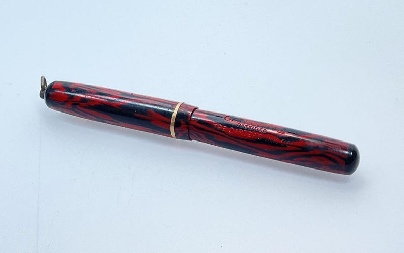 May include: A red and black marbled fountain pen with a gold band and a clip. The pen has the text "PATENTED" and "THE CONRAD CO. INC." on the side.