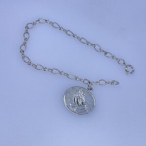 May include: Silver-toned bracelet featuring an oval charm with a detailed ship design. The bracelet has a delicate chain with an oval link pattern and a secure clasp. The charm depicts a sailing ship on the water.