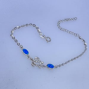 May include: A silver bracelet featuring a sea turtle charm and two oval blue opal stones. The bracelet has a delicate chain design and a lobster clasp closure. The bracelet is a jewelry item.