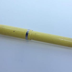 May include: A vintage yellow fountain pen with silver-colored accents. The pen has a cylindrical body and a cap on the end. The pen is lying on a white surface.