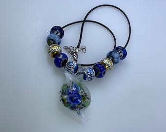 May include: A blue and white glass pendant necklace with a floral design, featuring a black cord and blue and gold beads.