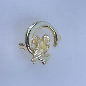 May include: A gold-colored metal pin featuring a cherub figure within a crescent-shaped design. The cherub is detailed with visible wings and facial features. The pin has a shiny, reflective surface.