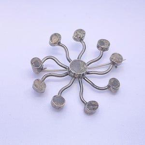 May include: A circular silver-toned brooch featuring a central round stone surrounded by nine radiating curved metal arms, each tipped with a small, matching round stone. The metal has a smooth finish, and the stones exhibit a mottled grey texture.