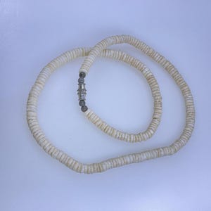 May include: A cream-colored necklace made of flat, round shell beads. The beads are strung together, forming a circular shape. The necklace is fastened with a silver-colored clasp. The necklace is displayed on a white surface.