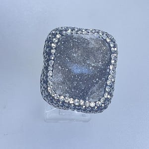 May include: A statement ring featuring a large, rectangular, gray druzy stone. The stone is surrounded by a border of small, clear crystals set in a silver-tone metal. The ring is displayed on a clear stand.