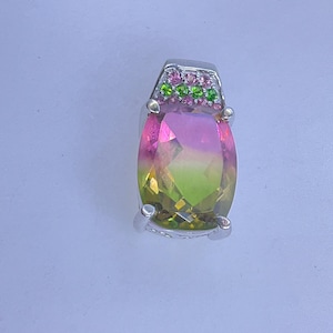 May include: A silver pendant featuring a large, oval-cut gemstone with a gradient of pink, yellow, and green hues. The top of the pendant is adorned with smaller pink and green gemstones, adding a touch of sparkle.