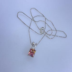May include: A silver necklace with a rectangular pendant featuring a multi-colored gemstone. The pendant is set with small, clear stones. The chain is a delicate box chain style.