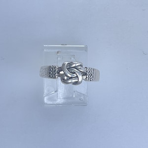 May include: A silver-toned ring featuring a knot design at the center. The ring has textured details on the band on either side of the knot. The ring is displayed on a clear, rectangular stand against a light blue background.