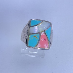 May include: A silver ring with a rectangular face featuring a mosaic design. The mosaic includes turquoise, pink, and white sections. The ring is displayed on a clear stand against a light background.