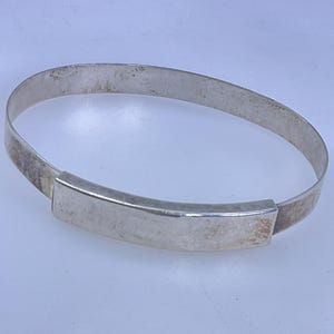 May include: A silver-toned metal bangle bracelet featuring a smooth, curved rectangular plaque centered on a thin, continuous band. The metal surface displays a polished finish with subtle signs of wear and light tarnishing across the band.