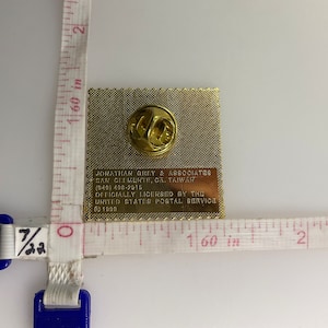 May include: A gold-colored pin with a detailed image of a stamp. The back of the pin has the text "Jonathan Grey & Associates,  Taipei, Taiwan (949) 400-2613 Officially Licensed by the United States Postal Service &copy; 1990".