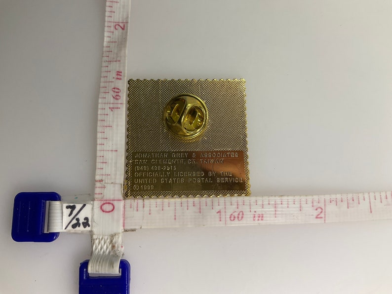 May include: A gold-colored pin with a detailed image of a stamp. The back of the pin has the text "Jonathan Grey & Associates,  Taipei, Taiwan (949) 400-2613 Officially Licensed by the United States Postal Service &copy; 1990".