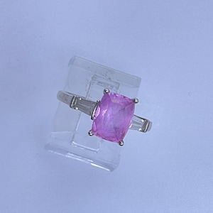 May include: A silver ring featuring a large, rectangular pink gemstone as the centerpiece. Two smaller, rectangular clear stones flank the main stone. The ring is displayed on a clear stand against a light blue background.