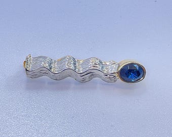 May include: A silver-toned brooch with a wavy design and a gold-toned oval setting holding a blue gemstone. The brooch has a clasp on one end.