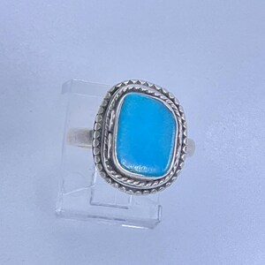 May include: A silver ring featuring a vibrant turquoise stone. The stone is rectangular with rounded edges, set within a textured, beaded silver frame. The ring is displayed on a clear stand against a light background.