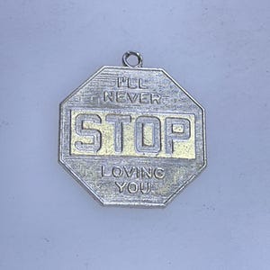 May include: Silver-toned octagonal pendant with the words "I'll Never Stop Loving You" in a gold-colored rectangle. The pendant has a textured surface and a small loop at the top for hanging.