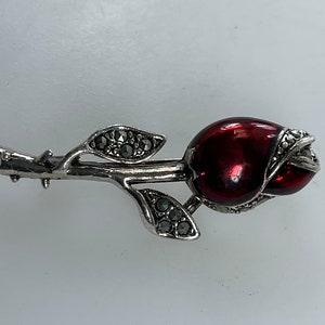May include: A silver rose brooch with a red enamel rose bud and marcasite accents.