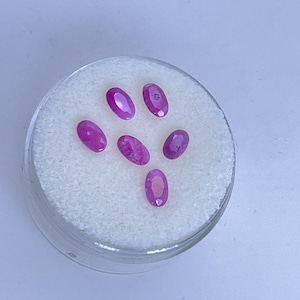 May include: A clear, round container holding six oval-shaped, vibrant pink gemstones. The gemstones are arranged on a bed of white, fluffy material. The container is set against a light background.