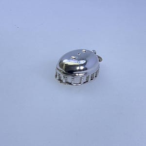 May include: A small, silver-colored charm in the shape of a bottle cap. The charm has a rounded top with several small indentations and a detailed, textured edge. It is designed to be a pendant or charm.