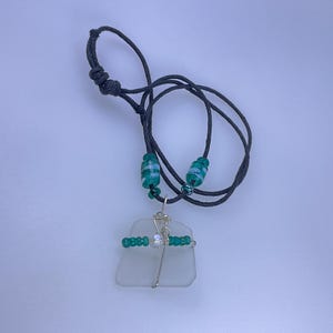 May include: A handmade necklace featuring a piece of sea glass wrapped in silver wire. The necklace has a black cord with green and white beads and a sliding knot closure. The sea glass is a light, frosted white color.