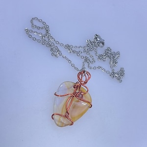 May include: A handcrafted necklace featuring a polished, heart-shaped stone pendant. The stone is a mix of cream and amber hues, wrapped in copper wire. The pendant hangs from a silver-colored chain, creating a unique and elegant accessory.