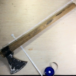 May include: A wooden-handled axe with a black metal head. The axe is lying on a wooden surface with a measuring tape next to it.