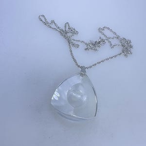 May include: A clear, triangular pendant necklace with a silver-colored chain. The pendant has a layered design with a spherical element inside. The necklace is displayed on a white surface, showcasing its reflective qualities.