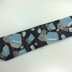 May include: A black tie with a repeating pattern of computer monitors, keyboards, and mice. The monitors are light blue with a white border. The keyboards are gray with black keys. The mice are gray with a black cord. The background is black with white dots.