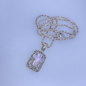 May include: A silver necklace with a pendant featuring a cross design. The pendant has a rectangular shape with an ornate, filigree border and a light purple center. The chain is a twisted rope style.