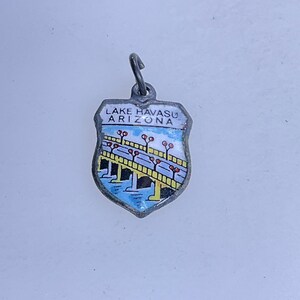 May include: A silver-toned shield-shaped charm with the words "Lake Havasu Arizona" in blue lettering. The charm features a bridge design with yellow, blue, and red accents, and a small loop for attachment.