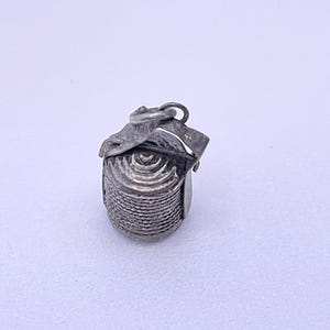May include: A silver-toned metal charm shaped like a spool of thread. The cylindrical object features a textured, coiled surface pattern resembling wound yarn or thread, topped with a small metal loop for attachment to a chain or bracelet.