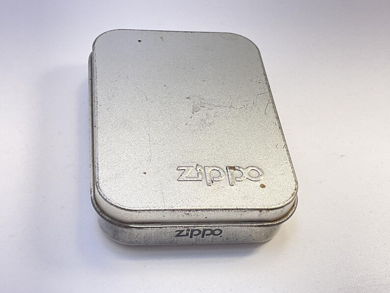 Puede incluir: Una caja met&aacute;lica rectangular de color plateado con bordes redondeados. La palabra "Zippo" est&aacute; grabada en la tapa y en la parte inferior de la caja. La caja tiene un aspecto ligeramente desgastado.