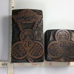 May include: Two antique printing blocks. The larger block is approximately 3 inches tall. Both blocks feature a clover design with text, including "Army of the Potomac" and "Association of the 22nd Brig."