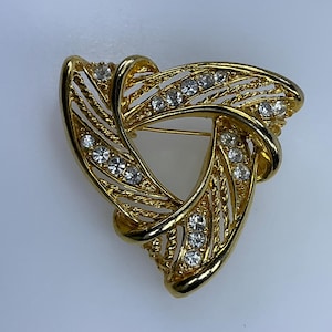 May include: A gold-toned brooch with a triangular shape, featuring a design of intertwined strands with clear rhinestones.