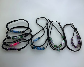May include: A set of five necklaces with black cords and colourful beads. The beads are made of clay and have a variety of patterns and colours, including blue, green, purple, and pink. Some of the necklaces have a small charm at the end.