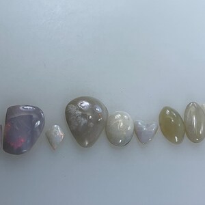 May include: A collection of eight iridescent opal stones in various shapes and sizes, including a heart, oval, and teardrop shapes. The stones are arranged in a row on a white surface.