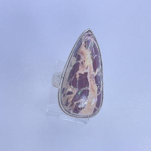 May include: A silver ring featuring a large, teardrop-shaped stone. The stone has a maroon base color with cream and tan veining. The ring is set on a clear display stand against a light blue background.