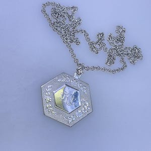 May include: A silver-toned necklace with a hexagonal pendant. The pendant features a central design with a gold-colored interior and a floral border. The chain is delicate and the pendant is detailed with engraved patterns.