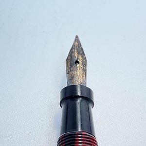 May include: Close-up of a black and gold fountain pen nib. The nib is pointed and has a small hole in the center.