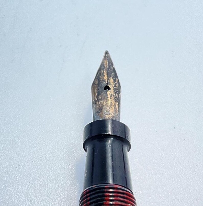 May include: Close-up of a black and gold fountain pen nib. The nib is pointed and has a small hole in the center.